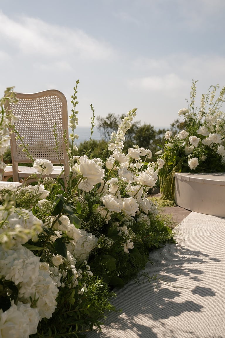 A sunny outdoor terrace decorated with white flowers in pots and a woven rattan chair under cloudy skies