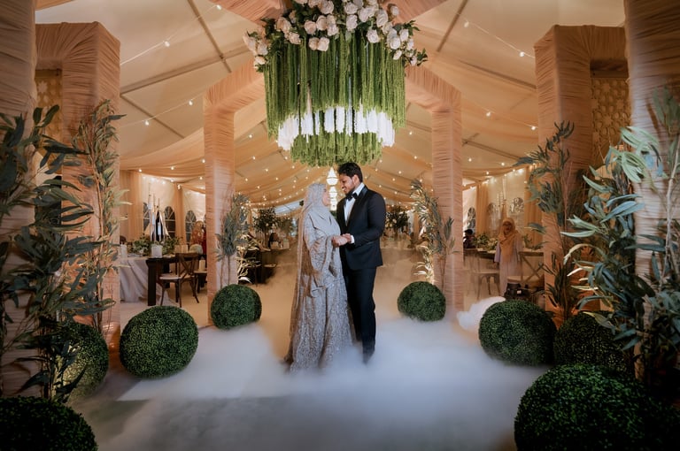 Bride and groom walking through an elegant venue with fog, hanging greenery, topiary plants, and warm lighting