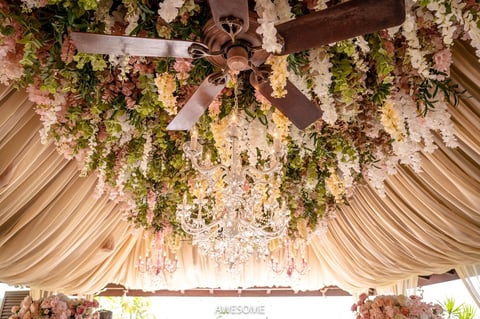 Elegant wedding tent ceiling with draped fabric, hanging greenery, flowers, and crystal chandelier creating a luxurious event space