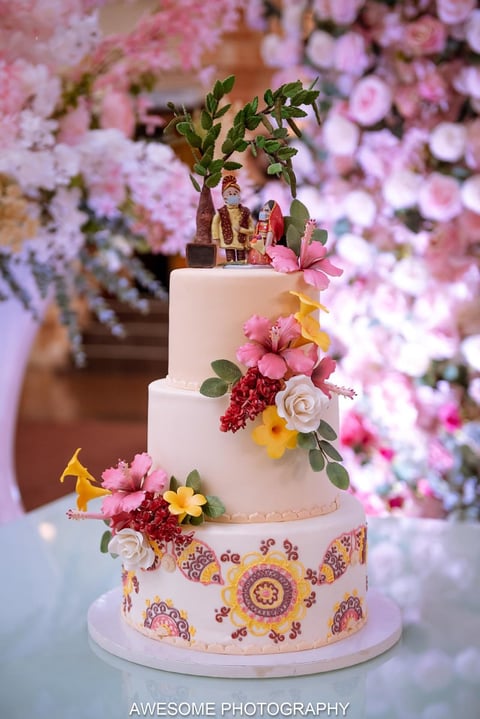Three-tiered white wedding cake with pink and yellow flowers, decorative patterns, and a bamboo plant topper, set against pink floral backdrop