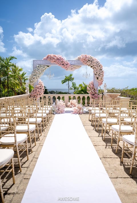 Elegant outdoor wedding ceremony setup with white aisle, pink floral arch, wooden chairs, and tropical palm trees under blue sky