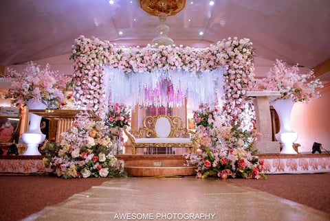 Elegant wedding stage decorated with abundant white and pink floral arrangements, ornate backdrop, chandelier, and dramatic purple lighting