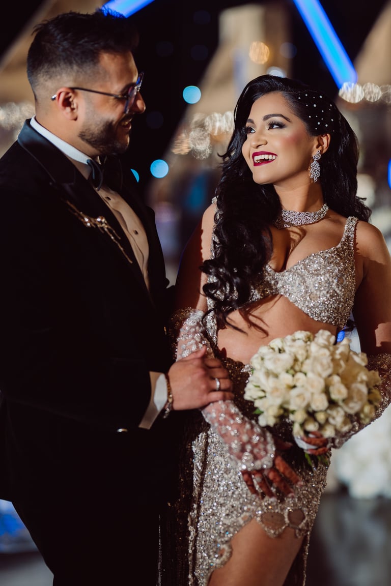 Elegant couple at formal event, woman in sparkly dress with white flower bouquet, man in black suit, nighttime outdoor setting with blue lights