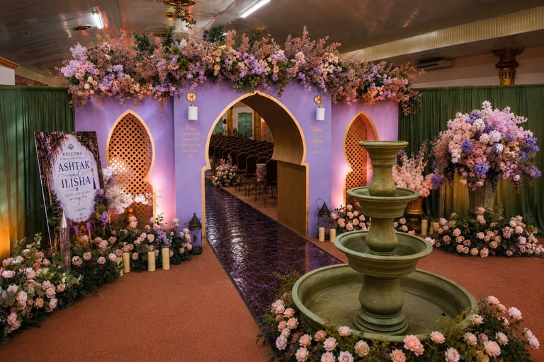 Ornate event venue decorated with purple lighting, white and pink flowers, archways, a central fountain, and candles on a red carpet