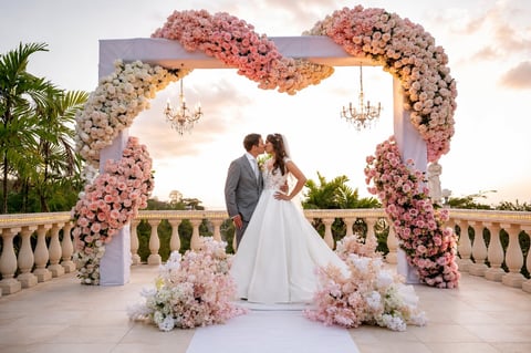 Bride and groom kissing under an ornate pink and white floral arch with hanging chandeliers at an outdoor wedding ceremony