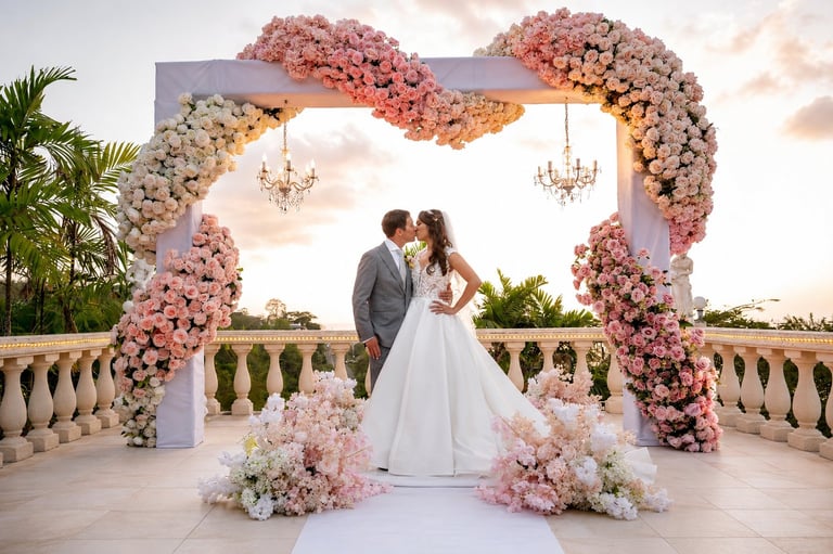 Bride and groom kissing under an ornate pink and white floral arch with hanging chandeliers at an outdoor wedding ceremony