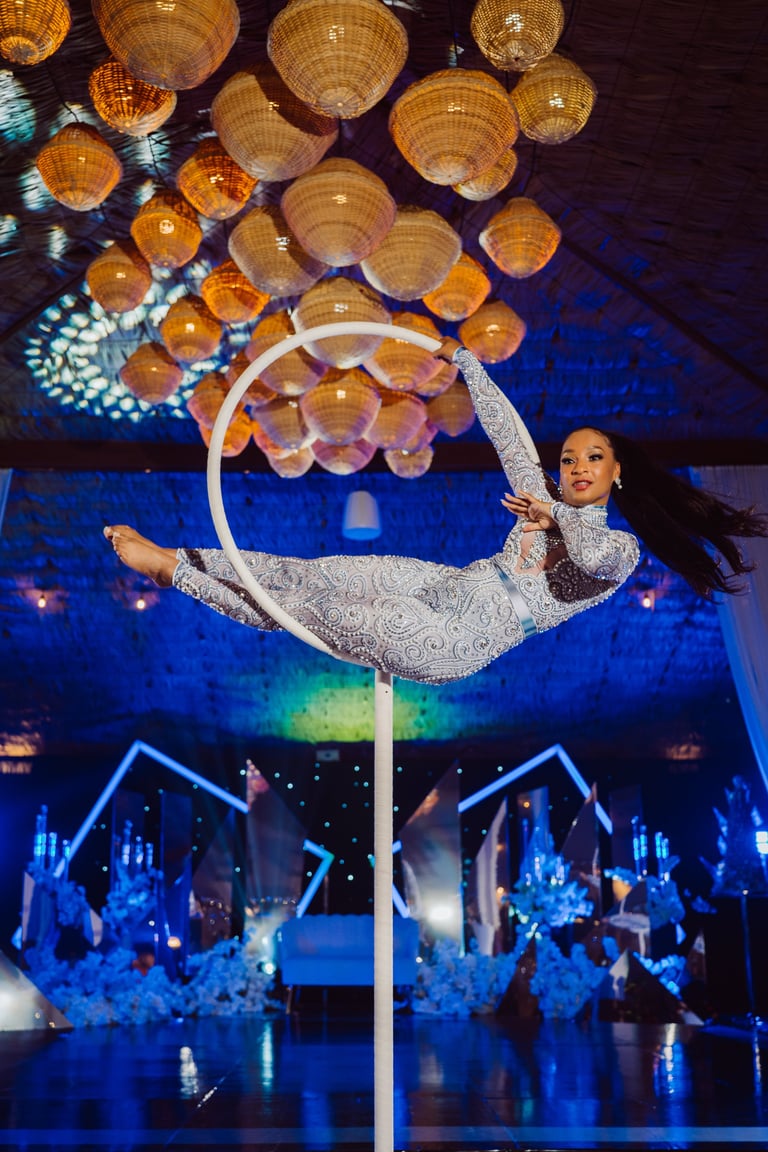Aerial performer in sequined outfit suspended on white hoop with woven pendant lights above in blue nightclub setting