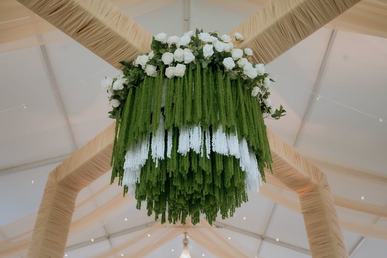 Hanging circular floral installation with green foliage and white flowers suspended from a white tent ceiling with wooden beams