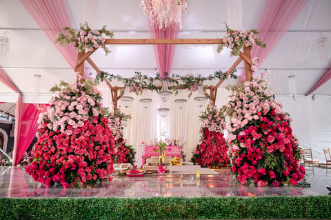 Elaborate wedding ceremony backdrop with pink and red floral arrangements, wooden arch, and draped pink fabric