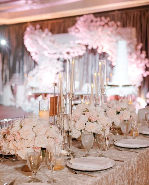 Elegant banquet table setting with cream-colored rose centerpieces, tall glass candelabras, white place settings, and pink balloon backdrop