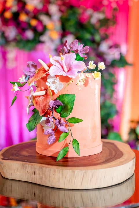 Elegant two-tiered cake with peachy-white frosting decorated with colorful edible flowers on wooden cake stands against vibrant pink and orange backdrop