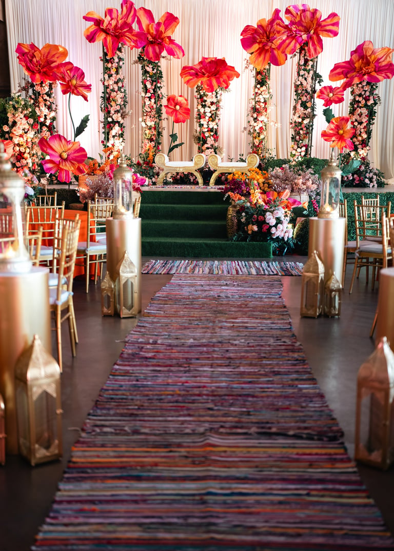 Luxurious event venue with vibrant red-pink floral installations lining both sides of a colorful striped aisle runner and cushioned seating