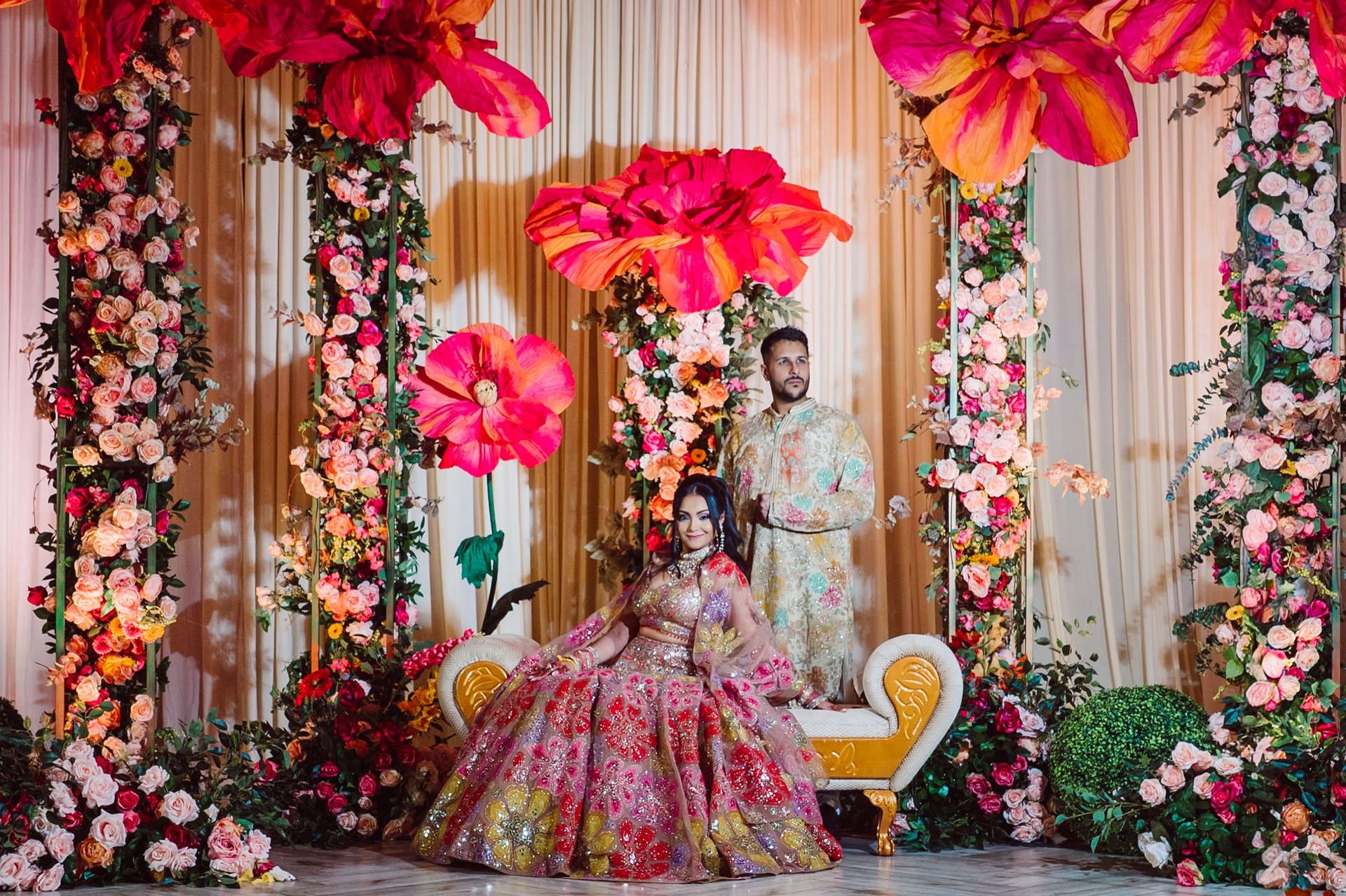 Couple posing in elaborate floral display with giant colorful flowers and hanging floral walls in wedding or event setting