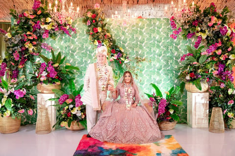 Bride and groom in traditional pink attire sitting under a floral arch with pink flowers and green foliage backdrop