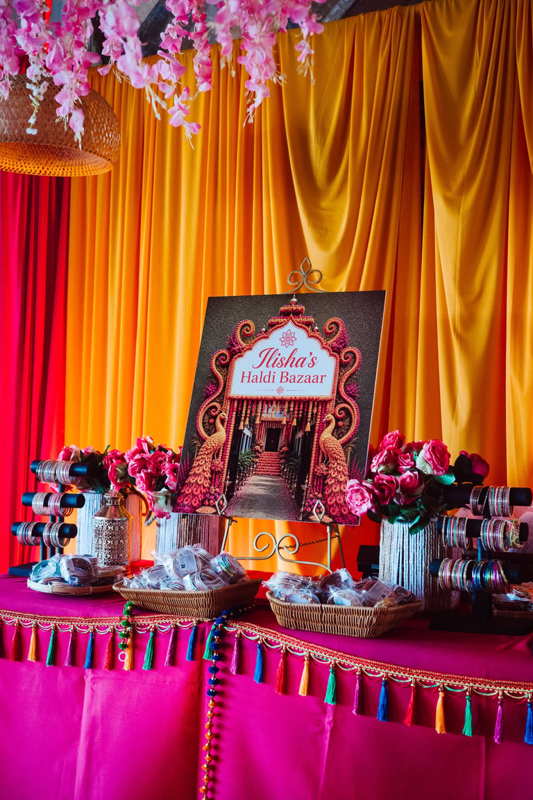 Colorful Indian wedding or event display with pink flowers, gold and magenta drapes, ornate signage reading Hishas Haldi Bazaar, and decorative bangles on a vibrant backdrop